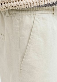 Gros plan sur un pantalon beige avec poche latérale et passants pour ceinture, associé à un pull en maille crème rentré.