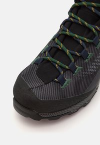 Chaussure de randonnée avec une tige grise et noire à motifs, des lacets verts et jaunes, et une semelle en caoutchouc noir robuste. Le tissu texturé améliore l'adhérence.