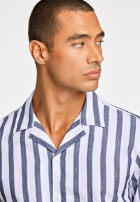 Linnen overhemd met verticale blauwe en witte strepen, korte kraag en knoopsluiting. De textuur lijkt licht en ademend.