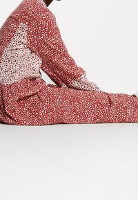 Långärmad jumpsuit i röd och vit prickig design, med en blandning av mönster och en texturerad kvalitet. Samlad midja och avslappnad passform.