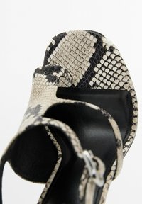 Svarta och beige sandaler med ormskalärmönster, med en texturerad överdel, justerbar spännesrem och en slät svart innersula. Unika mönsterdetaljer synliga.