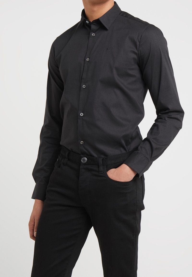 Camisa negra de botones con tela suave, que cuenta con mangas largas, un cuello clásico y un logotipo sutil en el pecho. Combinada con pantalones negros.
