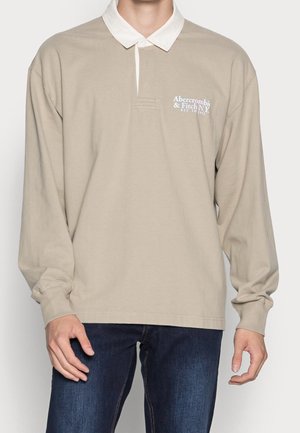 Langærmet beige polo t-shirt med hvid krave, der har diskrete syninger og et logo på venstre side. 100% bomuldsstof.