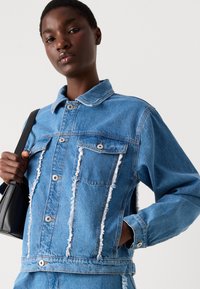 Giacca in denim blu con dettagli sfrangiati, due tasche frontali, chiusura con bottoni e colletto rialzato. Il materiale ha una superficie leggermente strutturata.