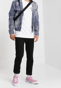 Blauwe en witte geprinte jas met rits over een wit T-shirt, gecombineerd met zwarte opgerolde broek en roze sneakers met witte veters.