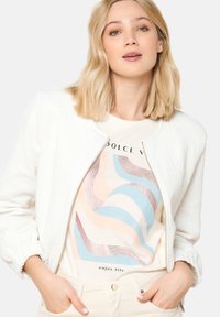 Bela struktura jakna s okroglim ovratnikom, z zadrgo, oblečena čez kremasto majico z pastelno valovito obliko in napisom. Kremaste hlače dopolnjujejo videz.