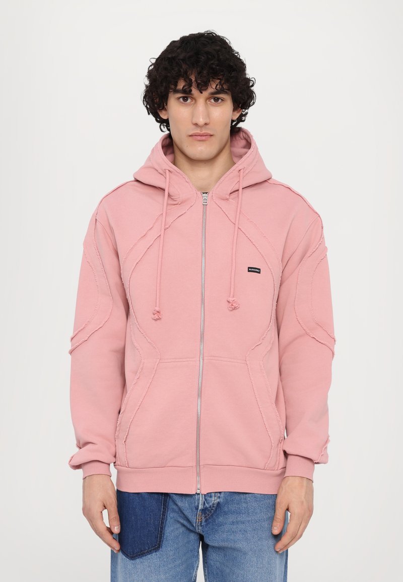 Rosafarbene Kapuzenjacke mit einem strukturierten Design, ausgestattet mit einer Kapuze, Zugbänder und kontrastierenden Nähten. Kombiniert mit blauen Jeans.