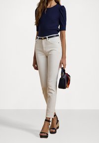 Žena v námořnickém topu, bílých vysokých džínách s černým opaskem, černých platformových sandálech, držící černou kabelku s oranžovým šátkem.