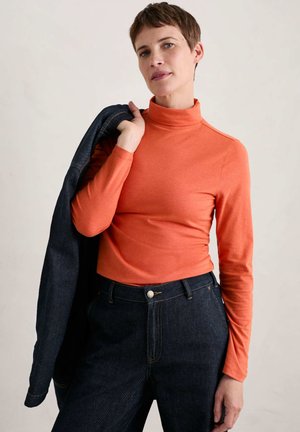 Oranje lange mouw turtleneck top gecombineerd met donkere spijkerbroeken; model houdt een blauwe jas vast. Glad materiaal, aansluitend ontwerp, verhoogde kraag.