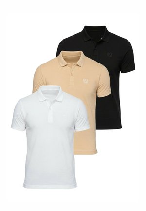 Homme portant un polo beige à manches courtes, un pantalon blanc large et des baskets blanches, debout devant un fond clair uni.
