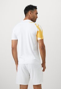 Witte sportshirt met gele gestreepte mouwen; korte mouwen, ronde halsontwerp en lichte stof. Draagt met bijpassende witte shorts.