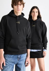 Čierne mikiny s vyšitým zlatým logom, klokanie vrecká, šnúrky na stiahnutie a rebrované manžety, v kombinácii so svetlomodrou košeľou a čiernou sukňou.