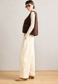 Brown fleece vest with two pockets, paired with cream wide-leg pants and brown heeled ankle boots, featuring ribbed long-sleeve top.