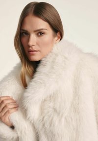 Biały płaszcz z sztucznego futra o grubej, miękkiej fakturze. Charakteryzuje się obszernym kołnierzem i warstwową konstrukcją. Model trzyma płaszcz na ramionach.