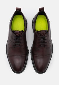 Une paire de chaussures en cuir marron foncé à lacets avec des semelles noires et des semelles intérieures jaunes vives, vue de dessus sur un fond blanc.