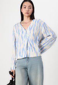 Knopfhemd mit langen Ärmeln in hellem Beige mit blauen vertikalen Streifen, V-Ausschnitt und lockerem Schnitt, kombiniert mit hellblauen Jeans.