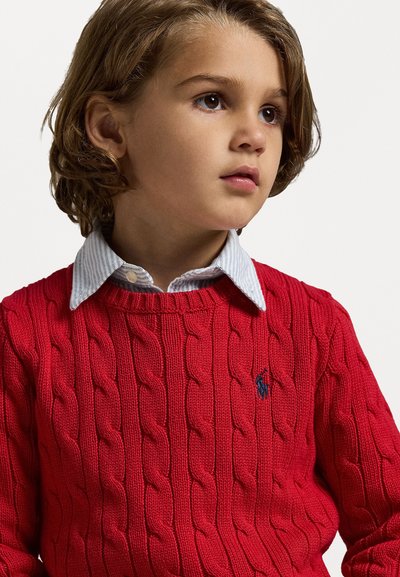 Roter Kabelstrickpullover aus Wolle mit Rundhalsausschnitt und navyfarbenem Logoakzent. Darunter ein blau-weiß gestreiftes Hemd mit Kragen.