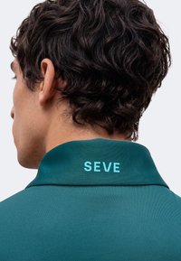 Jeune personne avec des cheveux courts, foncés et bouclés, portant une chemise bleu canard à col relevé, affichant le texte "SEVE" en bleu clair.