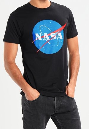 Mężczyzna ubrany w czarną koszulkę z niebieskim logo NASA z białymi literami i czerwonym znakiem, ręce w kieszeniach, czarne dżinsy.