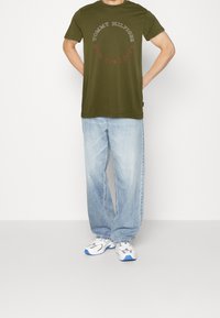 Camiseta de algodón verde oliva con mangas cortas y cuello redondo, que presenta un gráfico de "TOMMY HILFIGER NEW YORK CITY" en colores contrastantes. Llevado con jeans azul claro.