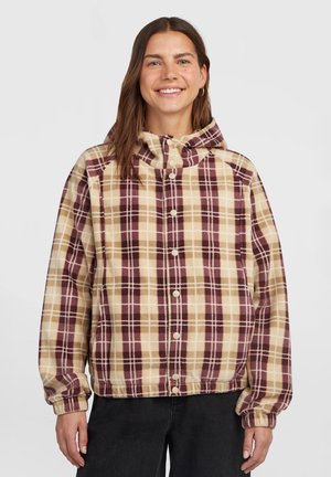 Lachende vrouw die een beige en bordeaux geruite jas met knopen, elastische manchetten en een capuchon draagt, staand tegen een effen achtergrond.