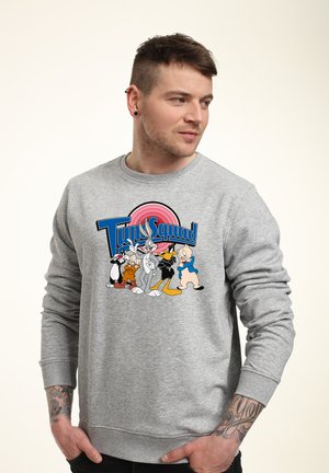 Hombre con sudadera gris con los personajes clásicos de Looney Tunes y texto "Tune Squad", mirando hacia la derecha con los antebrazos tatuados.