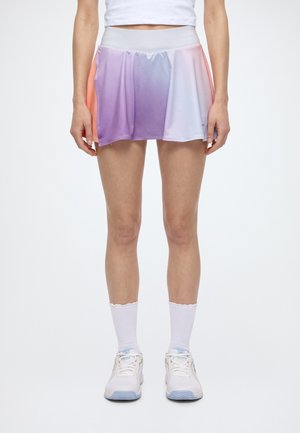 Modèle portant un crop top blanc, une jupe de sport violette pastel et blanche, des chaussettes blanches et des chaussures de tennis blanches.