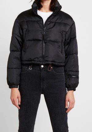 Person iført en kort sort dunjakke med høj krave, lynlås foran, sidelommer med lynlås og sorte højtaljede jeans.