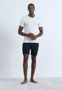 Bílé bavlněné tričko s kulatým výstřihem a malým logem, v kombinaci s černými šortkami. Model stojí bosý na světlém pozadí.