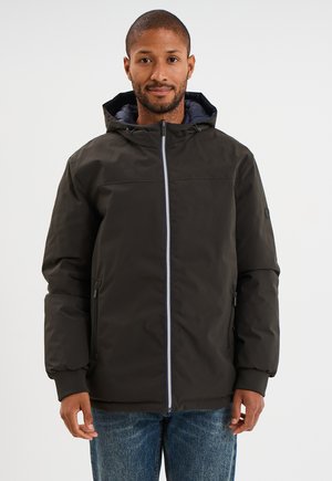 Hombre con chaqueta de invierno negra con capucha, cremallera y bolsillos laterales, combinada con vaqueros azules, de pie frente a un fondo blanco liso.