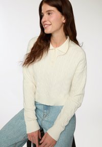 Donna con lunghi capelli castani che indossa un maglione bianco a treccia e jeans azzurri chiari, seduta e sorridente, guardando verso sinistra.