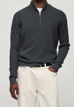 Homme portant un pull à demi-fermeture éclair côtelé gris foncé légèrement rentré dans un pantalon blanc cassé avec une ceinture noire, une main dans la poche.