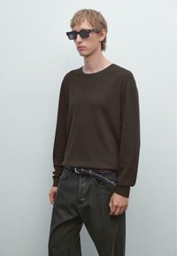 Junge Person mit kurzem blonden Haar, die dunkle rechteckige Sonnenbrillen, einen dunkelbraunen gerippten Pullover, schwarze Jeans und einen geflochtenen Gürtel trägt, vor einer schlichten Wand.