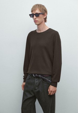 Young person with short blond hair wearing dark rectangular sunglasses, dark brown ribbed sweater, black jeans, and a braided belt against a plain wall.