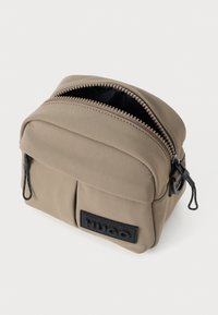 Tan, rektangulær kosmetiktaske med en tekstureret overflade, lynlås lukning og sort logo-patch. Indeholder to sidelommer og kontrasterende lynlås.