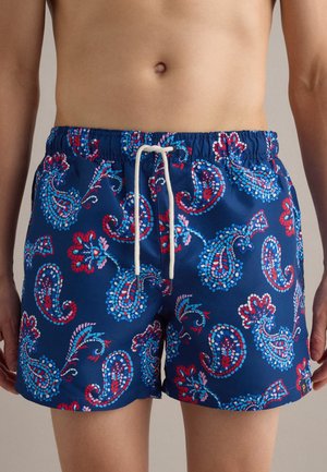 Männlicher Oberkörper mit blauen Badeshorts mit roten, pinken und hellblauen Paisley-Mustern und weißem Kordelzug.