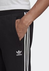 Pantaloni della tuta neri con tre strisce bianche sui lati. Presentano una vita regolabile e una tasca con zip. Logo Adidas in bianco.