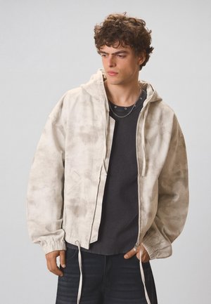 Young man with curly hair wearing a light beige zip-up hooded jacket over a black shirt, standing against a plain gray background.