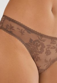 Nerifrån en persons höft med genomskinliga bruna spetsunderkläder med blommönster och vågiga kanter.