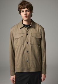 Olive green jacket made of textured fabric, featuring a button-up front, two chest pockets, and a curved hemline.
