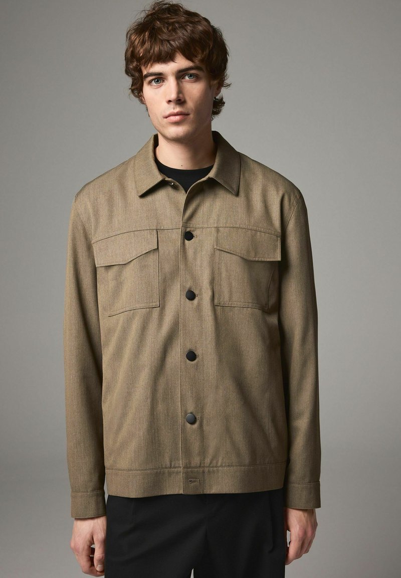 Olive green jacket made of textured fabric, featuring a button-up front, two chest pockets, and a curved hemline.