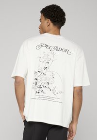 Valkea ylisuurikokoinen t-paita, jossa musta kukkakuva ja teksti "Pecador" selässä. Lyhyet hihat, rento istuvuus, puuvillamateriaali.