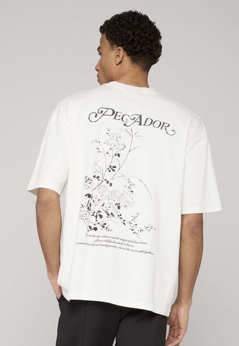 Valkea ylisuurikokoinen t-paita, jossa musta kukkakuva ja teksti "Pecador" selässä. Lyhyet hihat, rento istuvuus, puuvillamateriaali.
