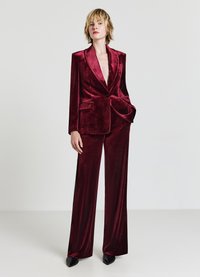 Burgunderroter Samtanzug mit Sakko mit spitz zulaufendem Revers, zwei Vordertaschen und weitem Bein bei der Hose, beide mit glatter, glänzender Textur.