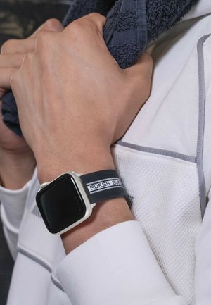 Crni gumeni remen za sat s bijelim logotipom "GUESS", srebrnim kućištem i pravokutnim crnim zaslonom. Nošen na ruci koja drži tamni ručnik.