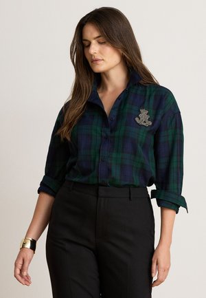 Femme portant une chemise à carreaux verte et bleu marine à boutons avec les manches retroussées, un pantalon noir et un bracelet en or au poignet gauche.