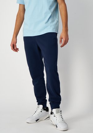 Mężczyzna w jasno niebieskim t-shircie, granatowych spodniach dresowych i białych wysokich trampkach stojący na jednolitym tle.