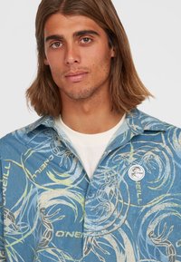 Camicetta a maniche corte con bottoni di colore blu, caratterizzata da un motivo onde tropicali, con accenti verde chiaro e una patch del logo O'Neill sul petto.