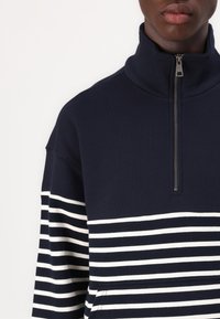 Navy zippered pullover with a high collar, featuring horizontal white stripes across the lower portion and a smooth texture.