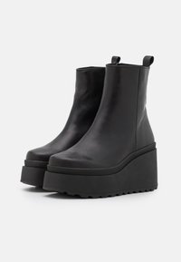 Bottines en cuir noir à plateforme avec une texture lisse, bout arrondi, semelle compensée haute, et deux tirettes en haut pour un enfilage facile.